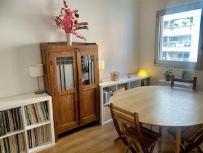A dining area with a round table, wooden chairs, a cabinet with glass doors, a lamp, and a window with a view of buildings. - Cozy Apartment in Central Paris (Paris)