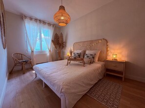Ein Schlafzimmer mit einem Bett, Nachttischen, einer Pendelleuchte, einem Sessel und einem Fenster mit Vorhängen.