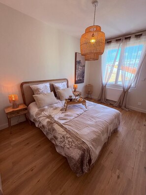 Ein Schlafzimmer mit einem großen Bett, einem Nachttisch mit Lampe, einer Pendelleuchte und einem Fenster mit Vorhängen.