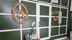 A green and white building facade with a door labeled 'HOME,' a wooden bench, a small table with a bottle and papers, and a decorative wreath.