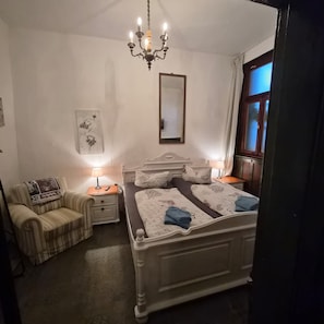 A bedroom with a four-poster bed, a nightstand with a lamp, a chair, and a window with curtains.