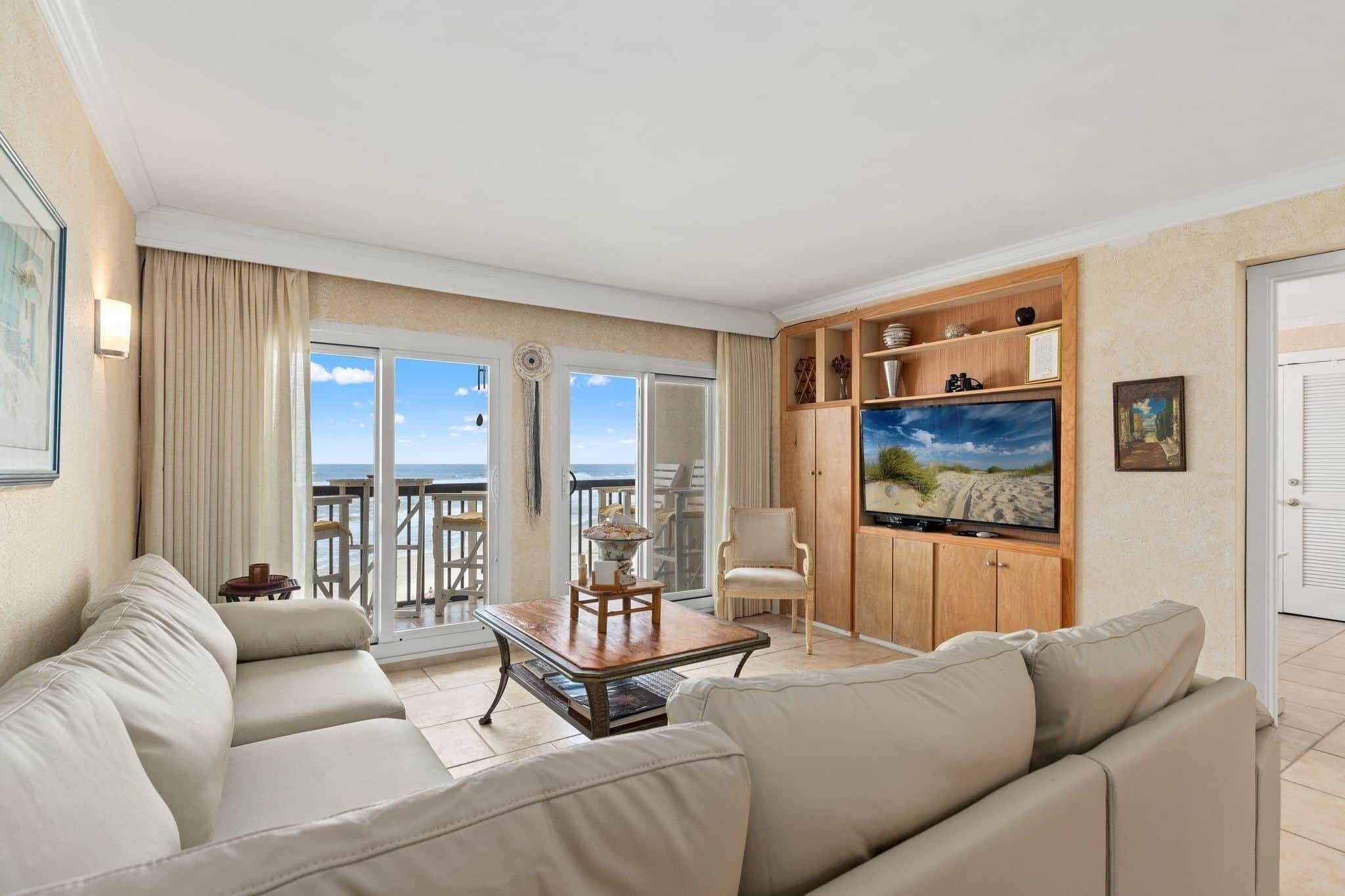 A spacious living room with a large TV, a sofa, a coffee table, and a balcony with a view of the beach.