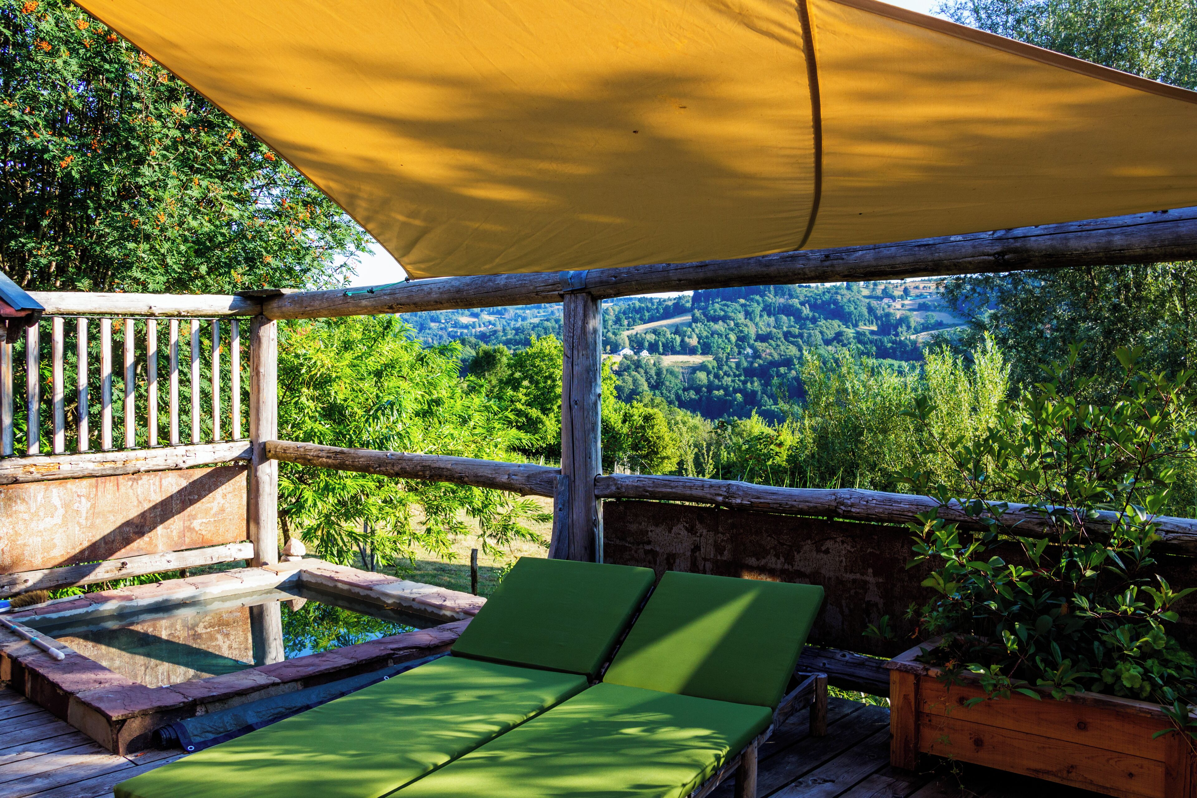 Een houten terras met een geel zonnescherm, groene loungestoelen en uitzicht op een bebost dal.