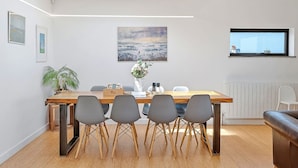 A modern dining area with a wooden table, four grey chairs, a vase with flowers, and a painting on the wall. - Bay House With Sea Views (Bude)