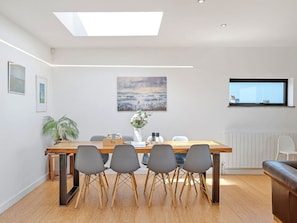 A modern dining area with a wooden table, four grey chairs, a vase with flowers, and a painting on the wall. - Bay House With Sea Views (Bude)