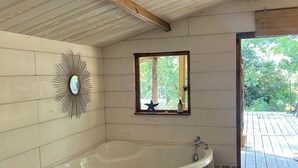 A bathroom with a freestanding bathtub, a wooden ceiling, a wall-mounted mirror, and a window with a view of greenery.