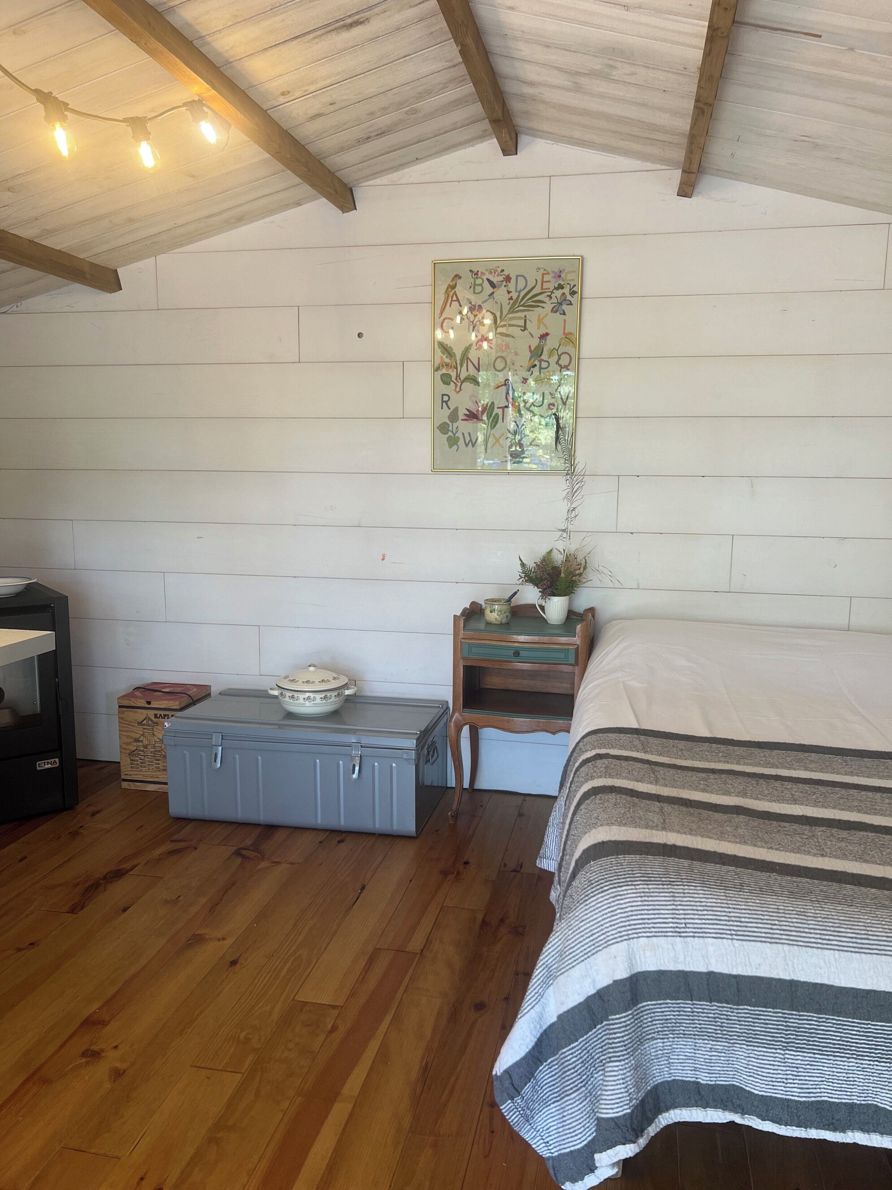 A bedroom with a wooden ceiling, a bed with a striped blanket, a nightstand with a plant, a wall-mounted painting, and a chest of drawers.