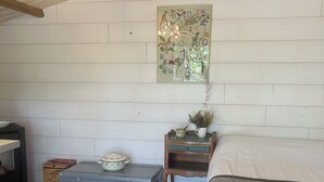 A bedroom with a wooden ceiling, a bed with a striped blanket, a nightstand with a plant, a wall-mounted painting, and a chest of drawers.