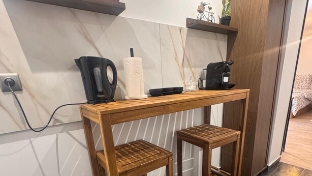 Et lille tekøkken med et træbord, to barstole, en kaffemaskine, en skraldespand og en hylde med røde beholdere.