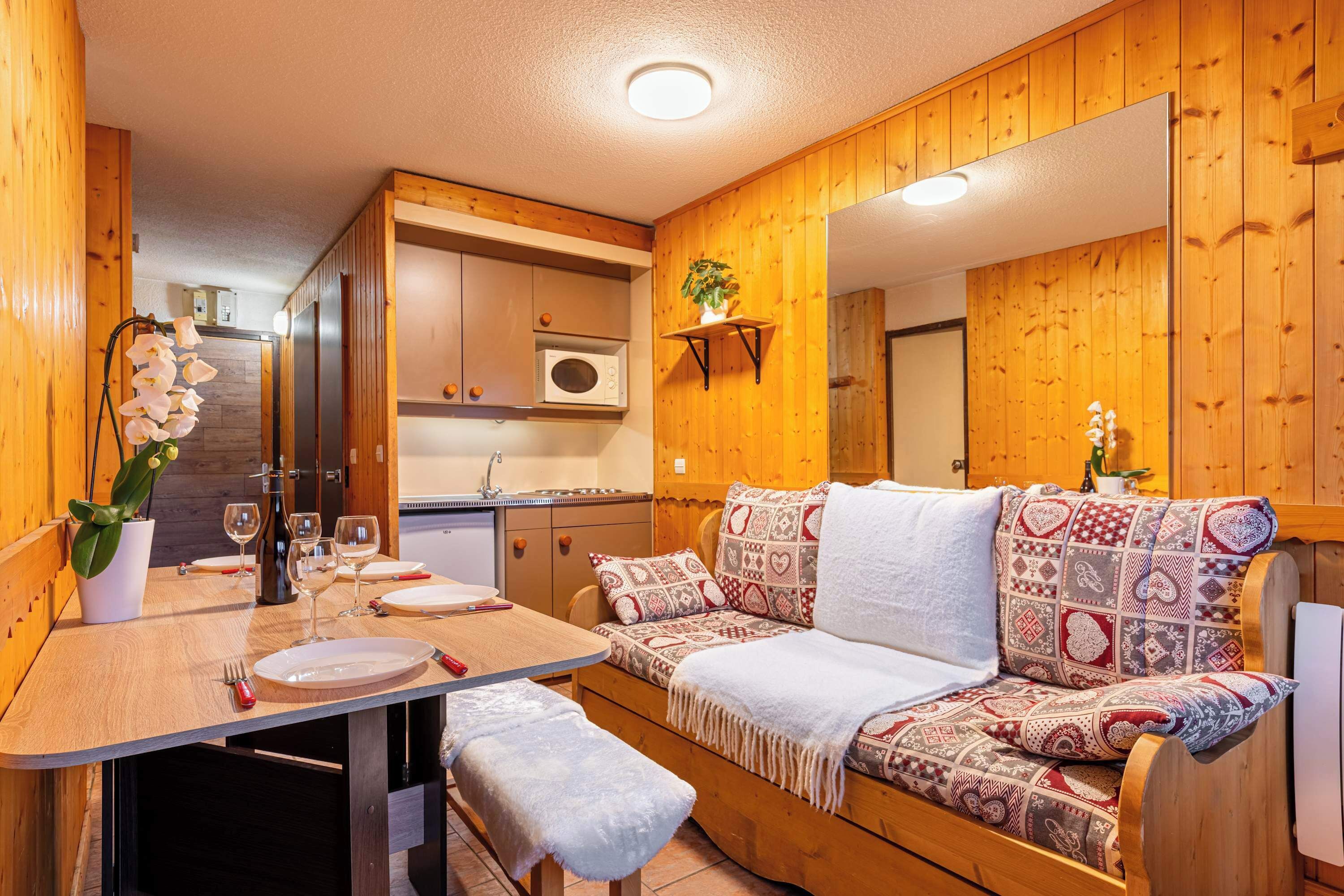 A cozy wooden interior with a dining table set for two, a sofa with patterned cushions, and a microwave in the kitchen area.