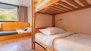 Ein Etagenbett mit Holz-Kopfteil und ein Einzelbett mit weißer Bettwäsche. Ein Fenster mit Vorhängen ist sichtbar, und durch das Fenster ist eine Landschaft zu sehen.