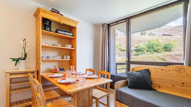 Ein gemütliches Zimmer mit einem Holzespeisebereich für vier Personen, einer Couch mit Kissen, einer Küchenzeile mit Geschirr und einem großen Fenster mit Blick ins Grüne.