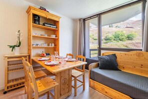 A cozy room with a wooden dining table set for four, a sofa with a cushion, a wooden cabinet with kitchen items, and a large window with a view of the countryside. - “V17” by Versant Collection (Les Belleville)