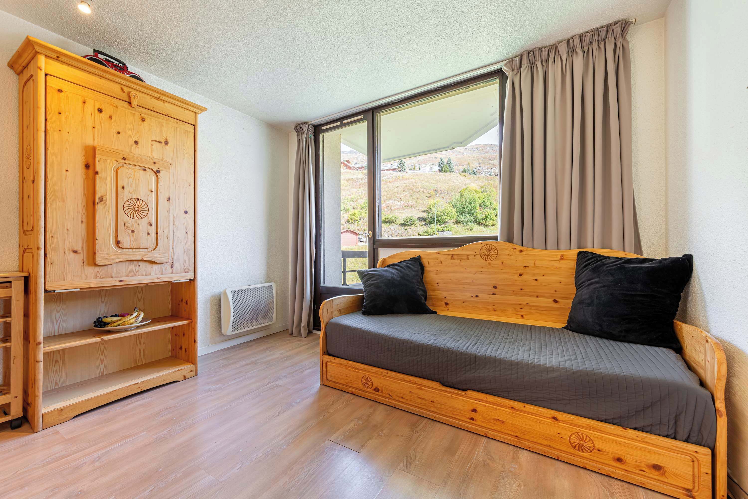 A wooden bed with a headboard, a wooden wardrobe with shelves, and a large window with curtains.