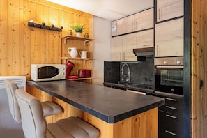 A compact kitchen with wooden cabinetry, a microwave, a red toaster, and a black countertop. - “V6” by Versant Collection (Val Thorens)