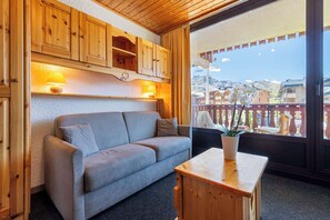 Ein gemütliches Zimmer mit einer grauen Couch, Holzschränken und einem großen Fenster mit Blick auf Gebäude und Berge.