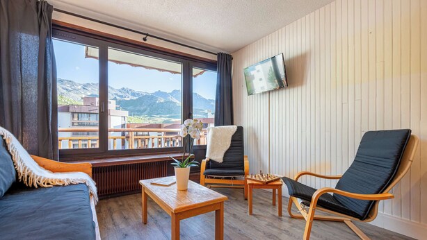 Una stanza con pavimento in legno, un divano, una poltrona, un tavolino con una pianta e un televisore fissato al muro.