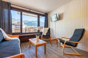 Um quarto com piso de madeira, um sofá, uma cadeira, uma pequena mesa com uma planta e uma TV fixada na parede.