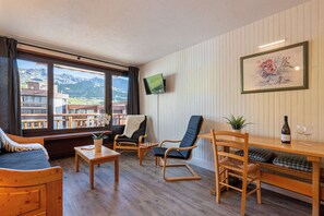 Ein gemütliches Zimmer mit einem Bett, einem Holztisch, zwei Stühlen, einem Fernseher und einem Fenster mit Blick auf die Berge.