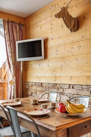 Ein Holzespeisetisch mit Tellern, Tassen und einer Obstschale. Über dem Tisch ist ein Fernseher an der Wand befestigt.