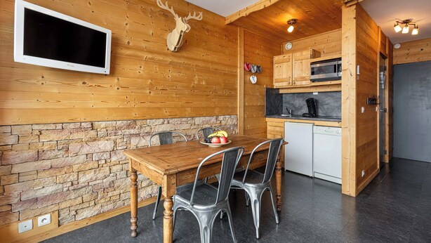Un interior de madera con una pared de piedra, un televisor montado, una mesa de comedor con sillas y una zona de cocina con armarios y electrodomésticos.