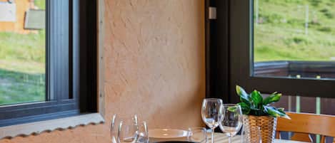 Mesa de comedor dispuesta con platos, tazones y copas de vino, con vistas a la vegetación a través de amplias ventanas.