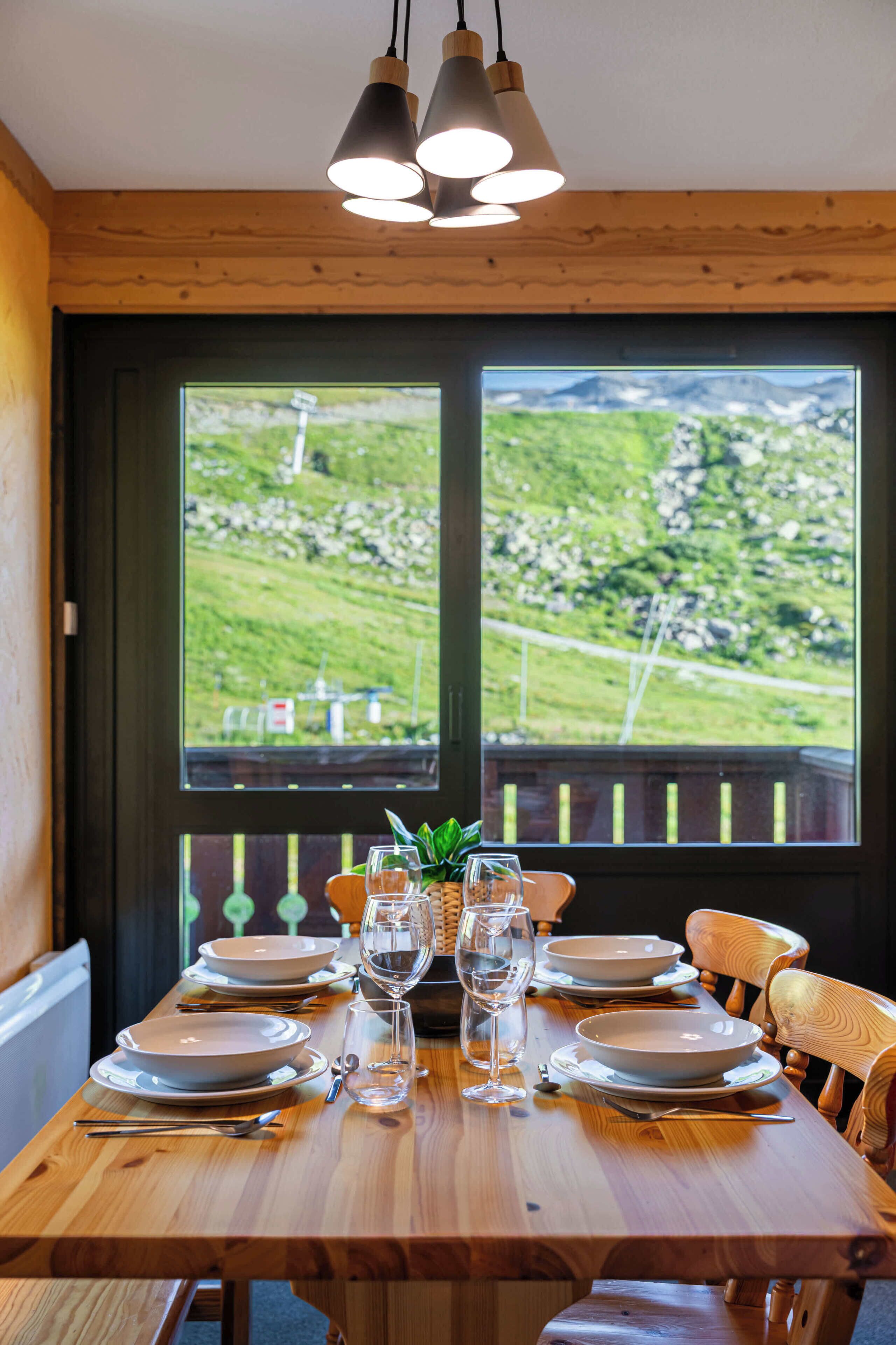 Une table à manger dressée avec des assiettes, des verres et des couverts, donnant sur un paysage verdoyant grâce à une grande fenêtre.
