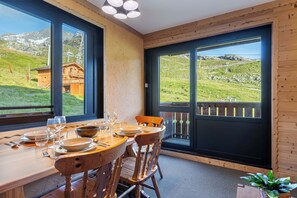 A dining area with a table set for four, large windows offering a view of a mountainous landscape, and a wooden interior. - Residence Olympic, Ski In-Out with WiFi in Val Thorens, “V11” by Versant Collection (Val Thorens)
