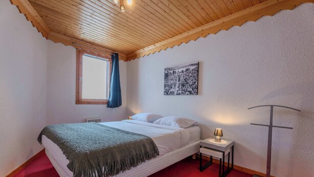 Ein Schlafzimmer mit Bett, Nachttisch, Lampe, einem Fenster mit Vorhang und einem Wandbild.