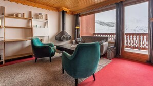 Ein gemütliches Zimmer mit Holzbalkendecke, Kamin, Sofa, zwei Sesseln, einem Tisch und Blick auf einen verschneiten Berg.