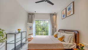 A bedroom with a bed, a bedside table, a nightstand, a ceiling fan, a window with curtains, and a wall-mounted shelf.