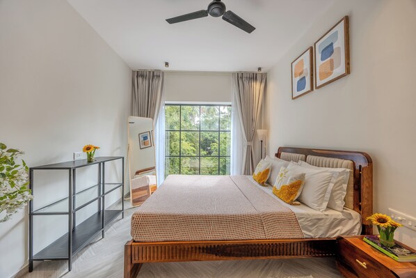 A bedroom with a bed, a bedside table, a nightstand, a ceiling fan, a window with curtains, and a wall-mounted shelf.