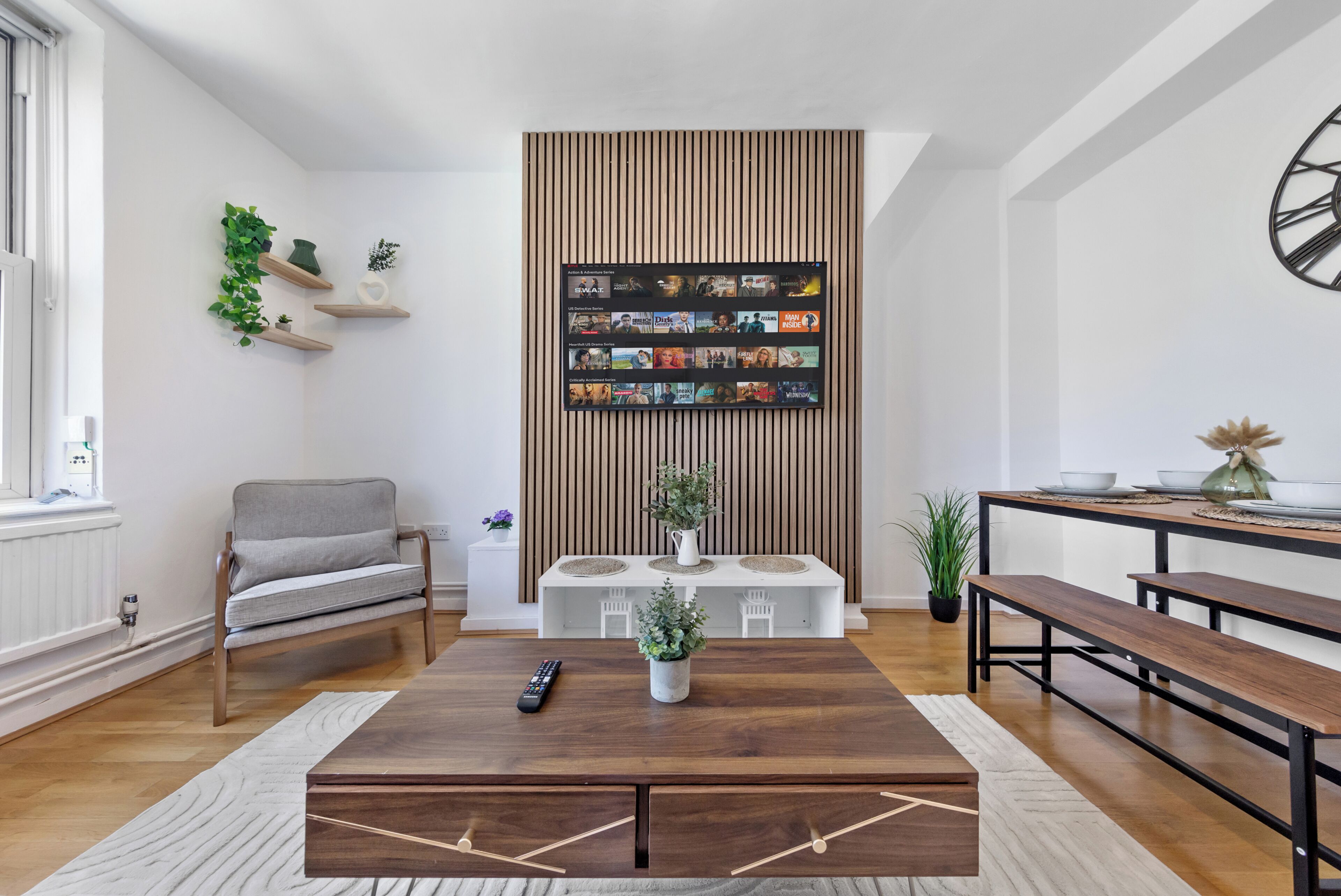 Ein Wohnzimmer mit Holztisch, einer Couch, einer TV-Wand mit Streaming-Diensten und einem Essbereich mit Bänken.