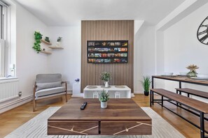 A living room with a wooden coffee table, a sofa, a TV wall with a streaming service interface, and a dining area with benches. - Spacious & Cosy Camden Town 3BR Sleeps 7! Only 10 Min to St Pancras Station! (London)
