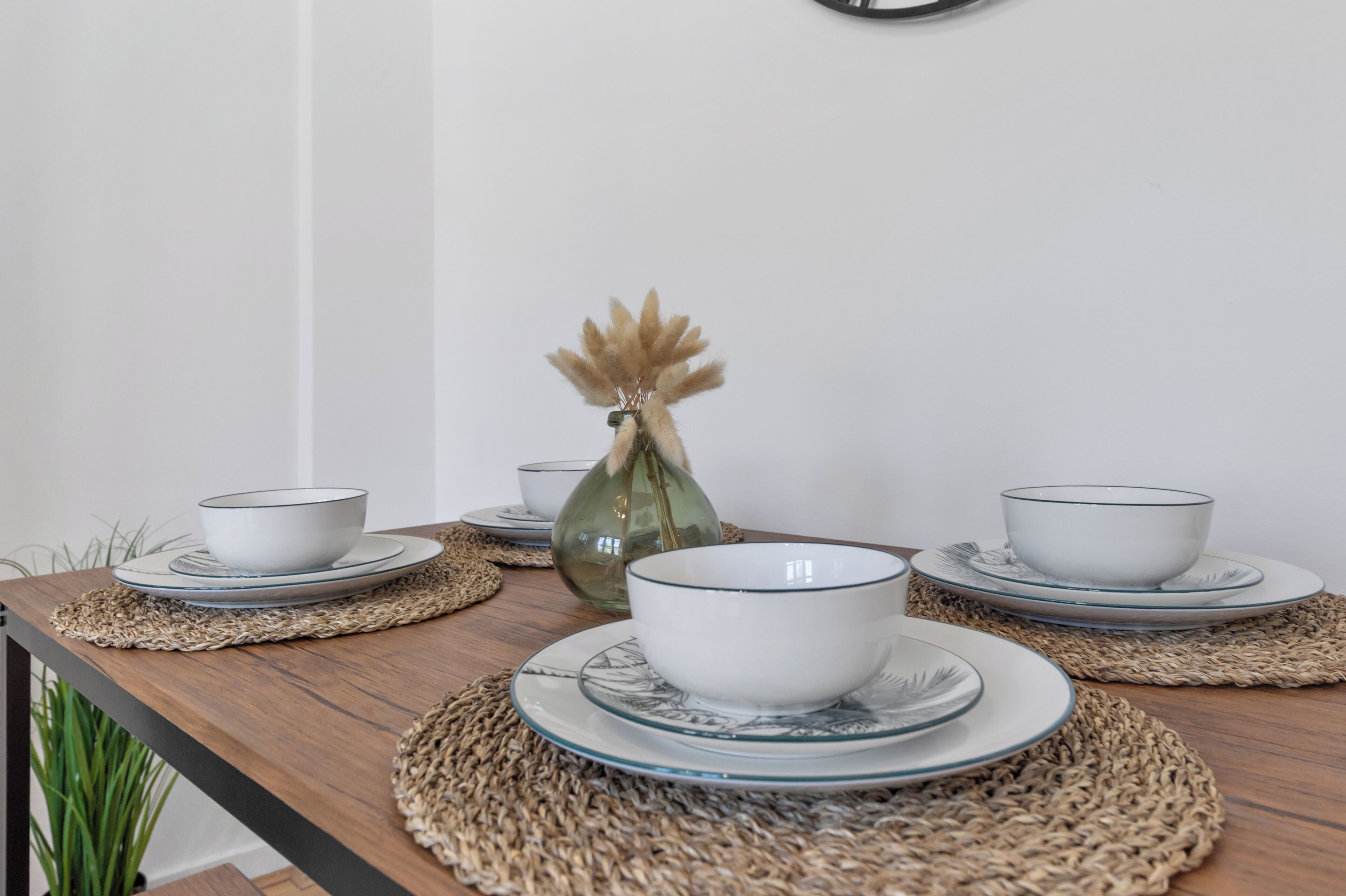 Una mesa de comedor con platos y tazones blancos, un florero de vidrio con flores secas y un mantel tejido.