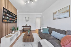 Ein modernes Wohnzimmer mit einem an einer Holzwand montierten Flachbildfernseher, einem Essbereich mit Tisch und Bänken, einem grauen Sofa mit Kissen und einem großen abstrakten Gemälde an der Wand.
