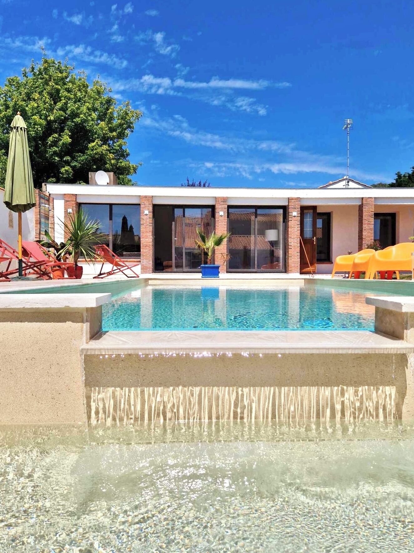 A swimming pool with a waterfall feature, surrounded by a building with large windows and a patio area with chairs and umbrellas.