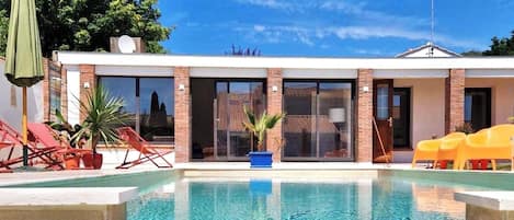 A swimming pool with a waterfall feature, surrounded by a building with large windows and a patio area with chairs and umbrellas.