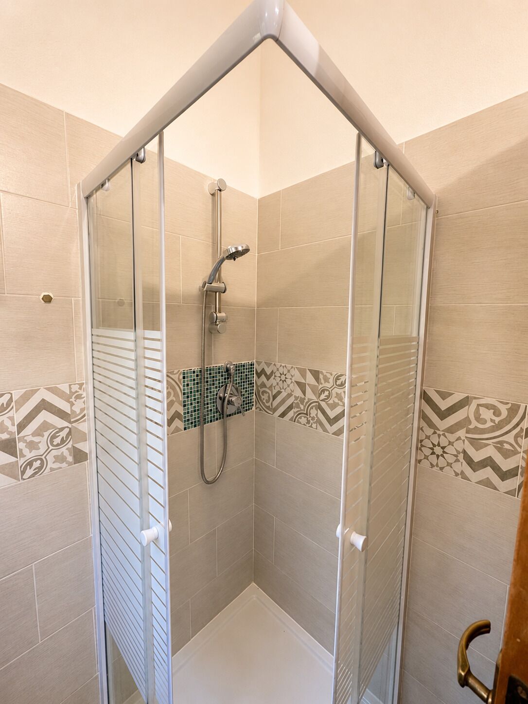 Um box de chuveiro com paredes revestidas de azulejos com padrões geométricos, porta de canto e chuveiro manual.