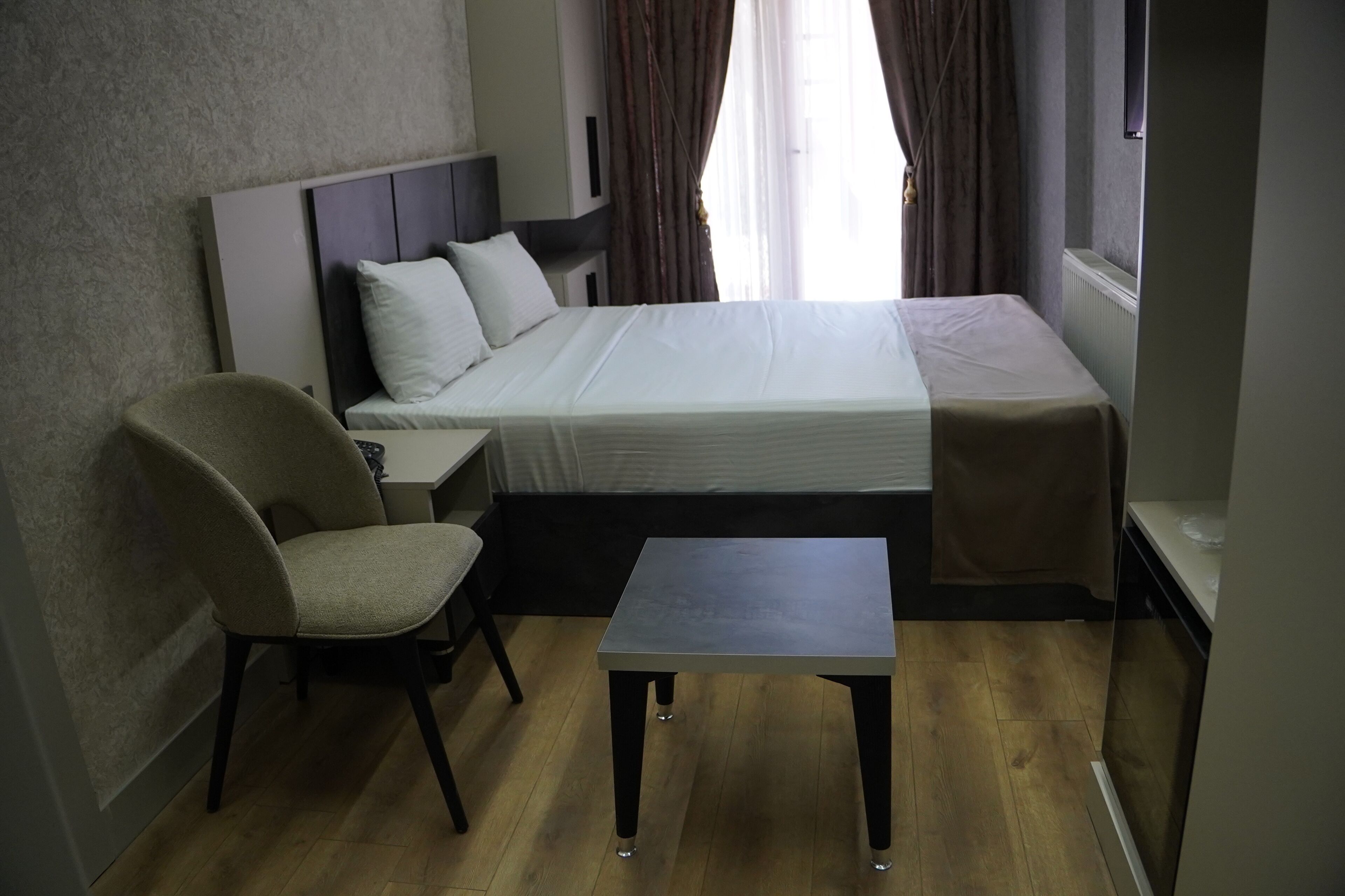 A hotel room with a bed, a chair, a small table, and a window with curtains.