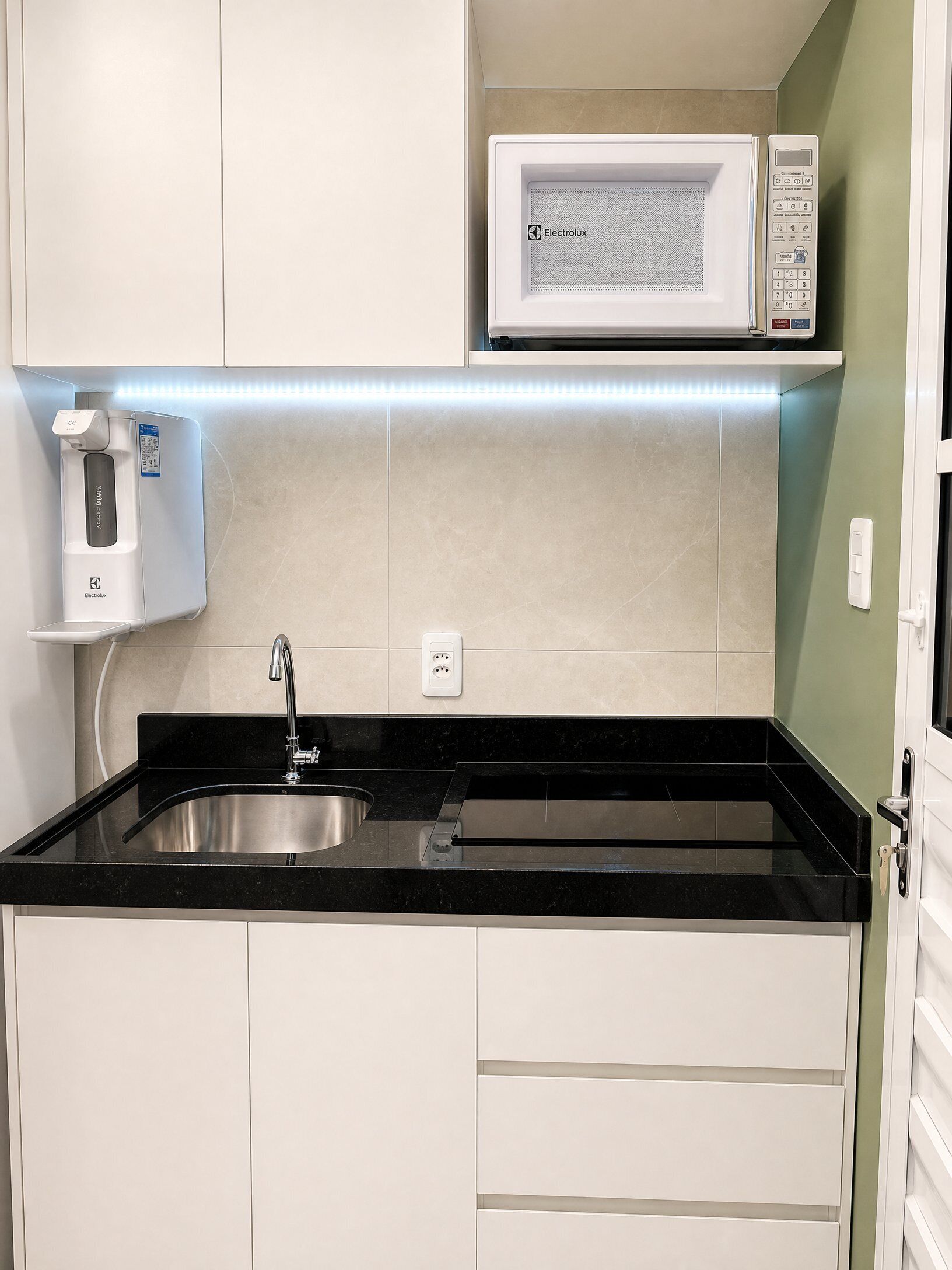 Una cocina con encimera negra, pileta de acero inoxidable, muebles blancos, microondas y dispensador de agua.