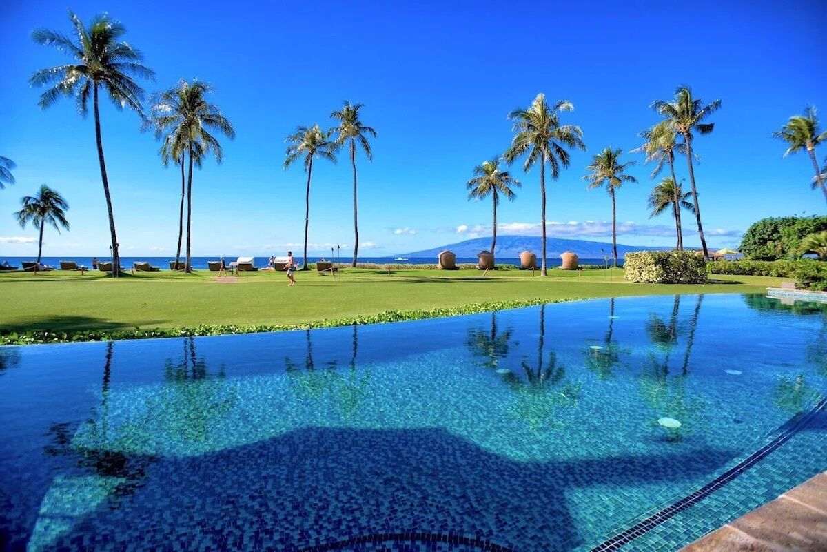 A clear blue infinity pool with a view of a golf course and palm trees.
