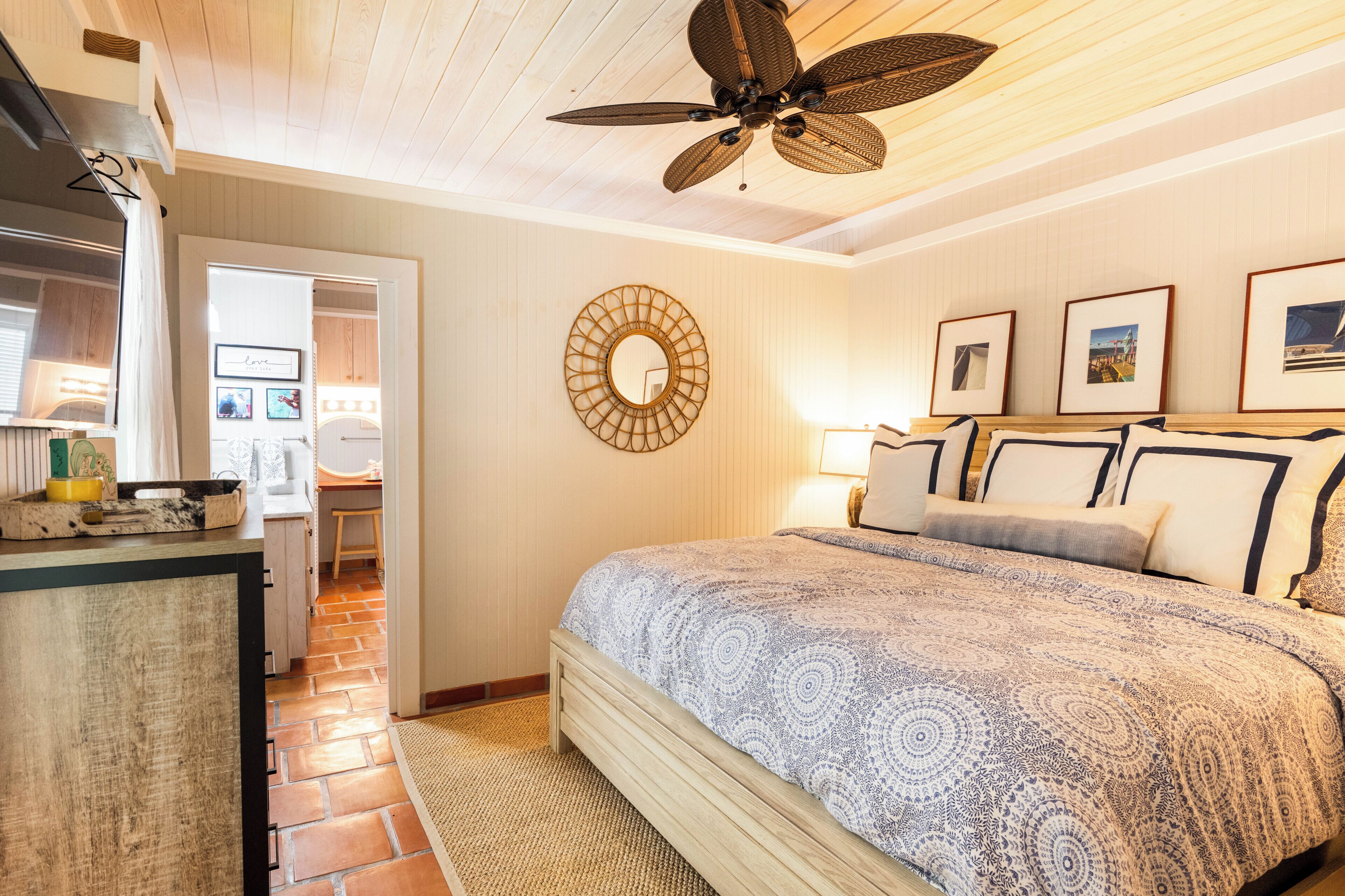 A bedroom with a bed, a ceiling fan, a wall-mounted mirror, and framed pictures.