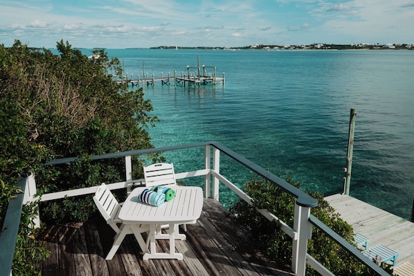 A wooden deck with a white chair and a table, overlooking a clear blue sea and a distant shoreline with buildings.
