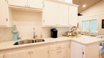 Una cocina con armarios blancos, un lavamanos, una cafetera y una zona de comedor con una mesa y sillas.