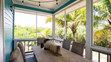A covered porch with wicker furniture, a wooden table, and a view of tropical vegetation.