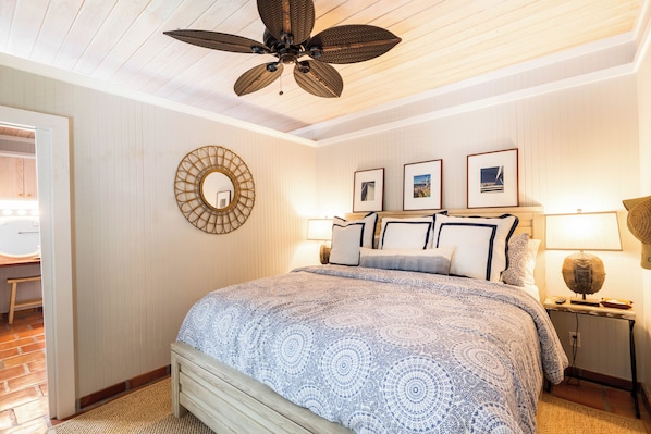 A bedroom with a large bed, a ceiling fan, a round mirror, and framed pictures on the wall.
