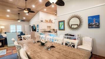 A dining area with a wooden table, white chairs, ceiling fans, and a kitchen in the background.