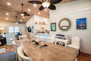A dining area with a wooden table, white chairs, ceiling fans, and a kitchen in the background.