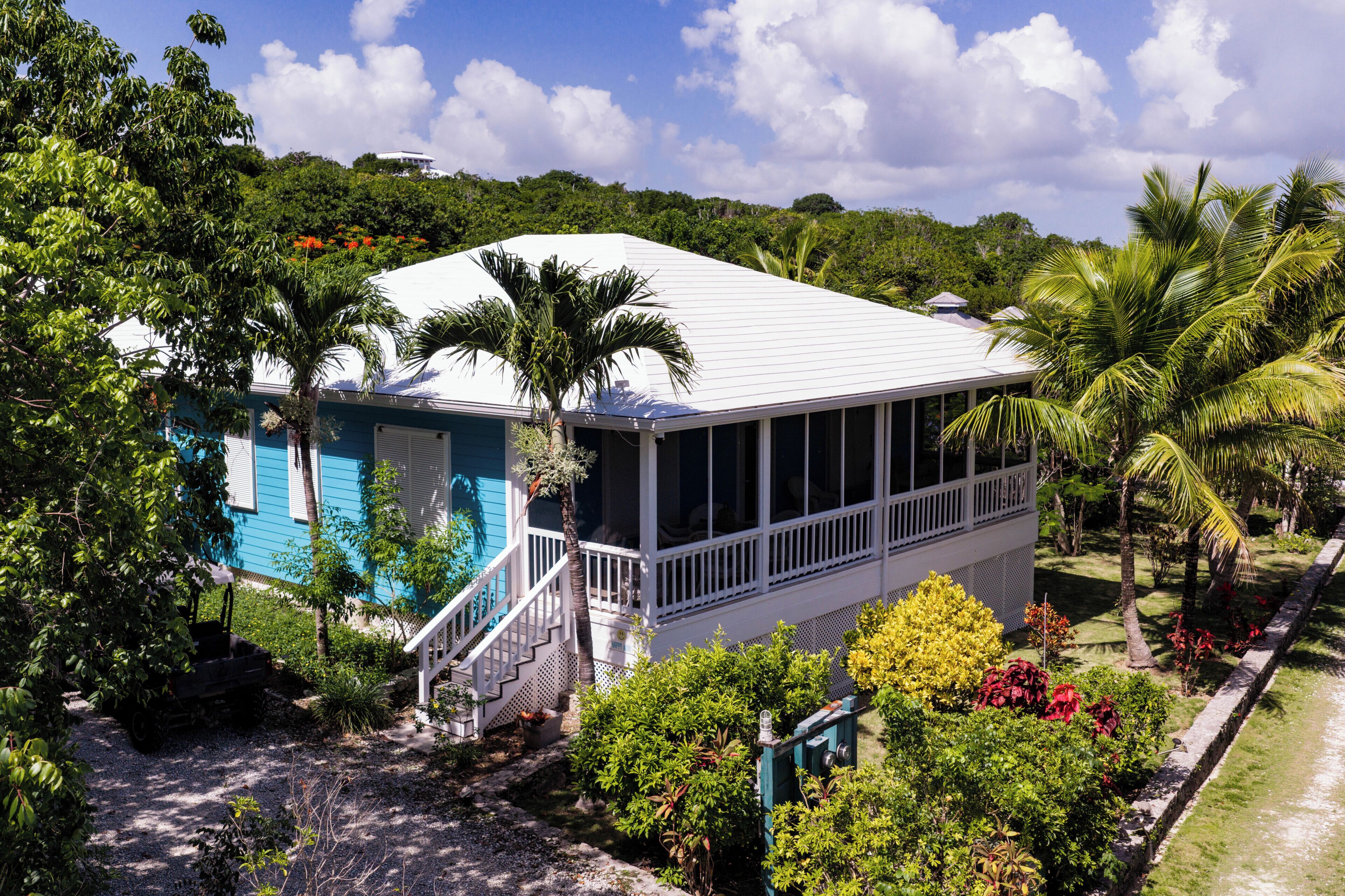 A two-story house with a white roof, surrounded by lush greenery and palm trees.